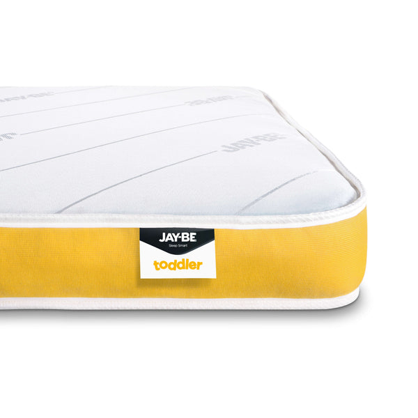 A close-up of a toddler mattress corner showing a white quilted top, a bright yellow side bolster, and a black and white Jay-Be toddler brand label.