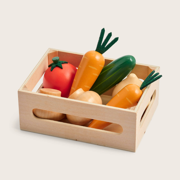 A wooden toy vegetable crate filled with colorful wooden play vegetables, including carrots, a tomato, a cucumber, and onions, on a light beige background.