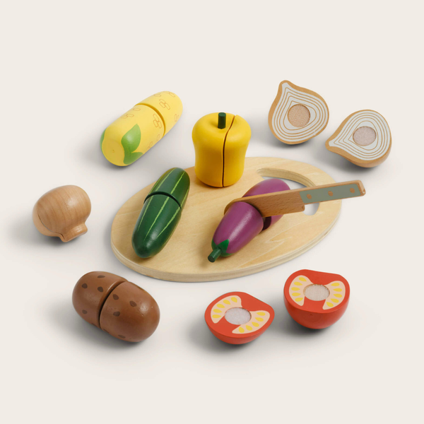 A wooden pretend-play kitchen vegetable and fruit cutting board set on a light beige background. The set includes a round wooden board, a small wooden knife, and various painted wooden vegetables and fruits such as a green cucumber, purple eggplant, yellow pepper, red tomato, brown potato, and sliced citrus.